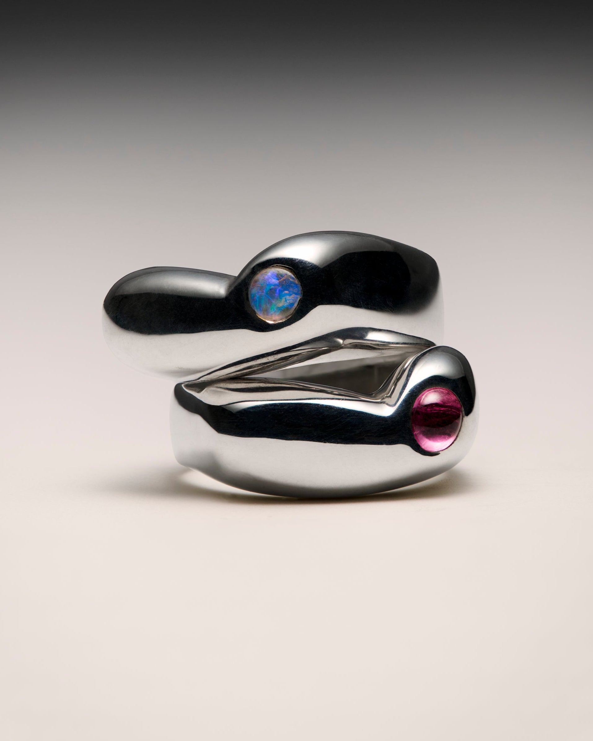 Two sculptural silver rings with organic, fluid shapes, one featuring a blue iridescent stone and the other a pink stone, displayed on a neutral gradient background.