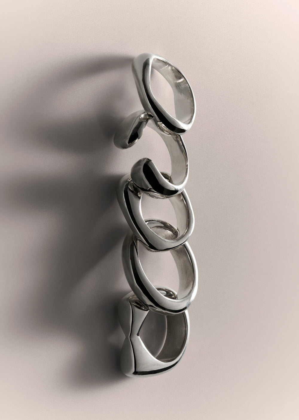 Four polished silver rings with organic, flowing curves arranged in a row on a warm beige background with soft shadows.