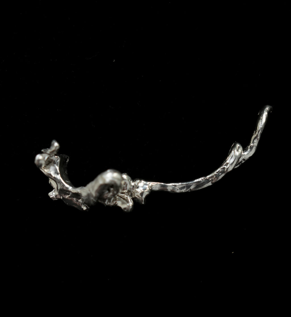 A single silver abstract sculptural object or jewelry piece with a textured organic curved form, photographed against a solid black background.