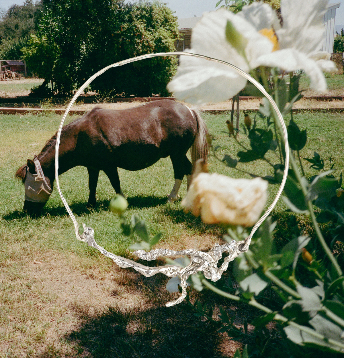 A silver-toned abstract sculptural ring pendant is shown hanging in front of a real outdoor scene with a brown cow grazing in a grassy field and white flowers in the foreground.