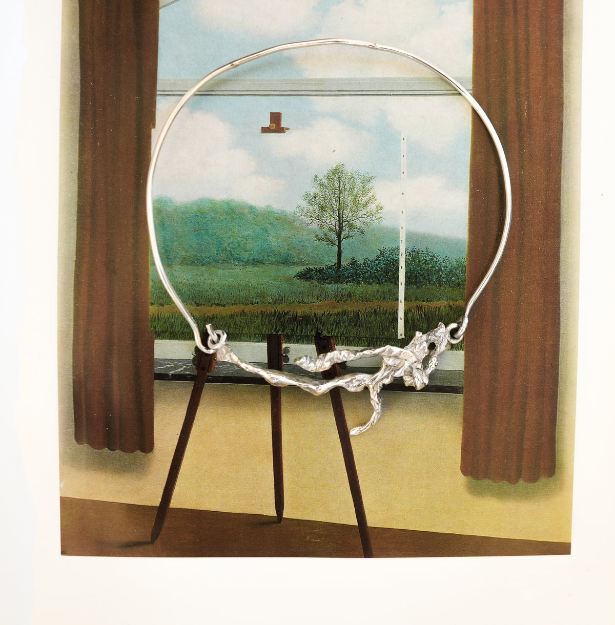 A silver collar necklace with organic shaped torque closure on top of a surreal art print or painting featuring a on a tripod easel, set against a window view of a green landscape with trees and brown curtains.
