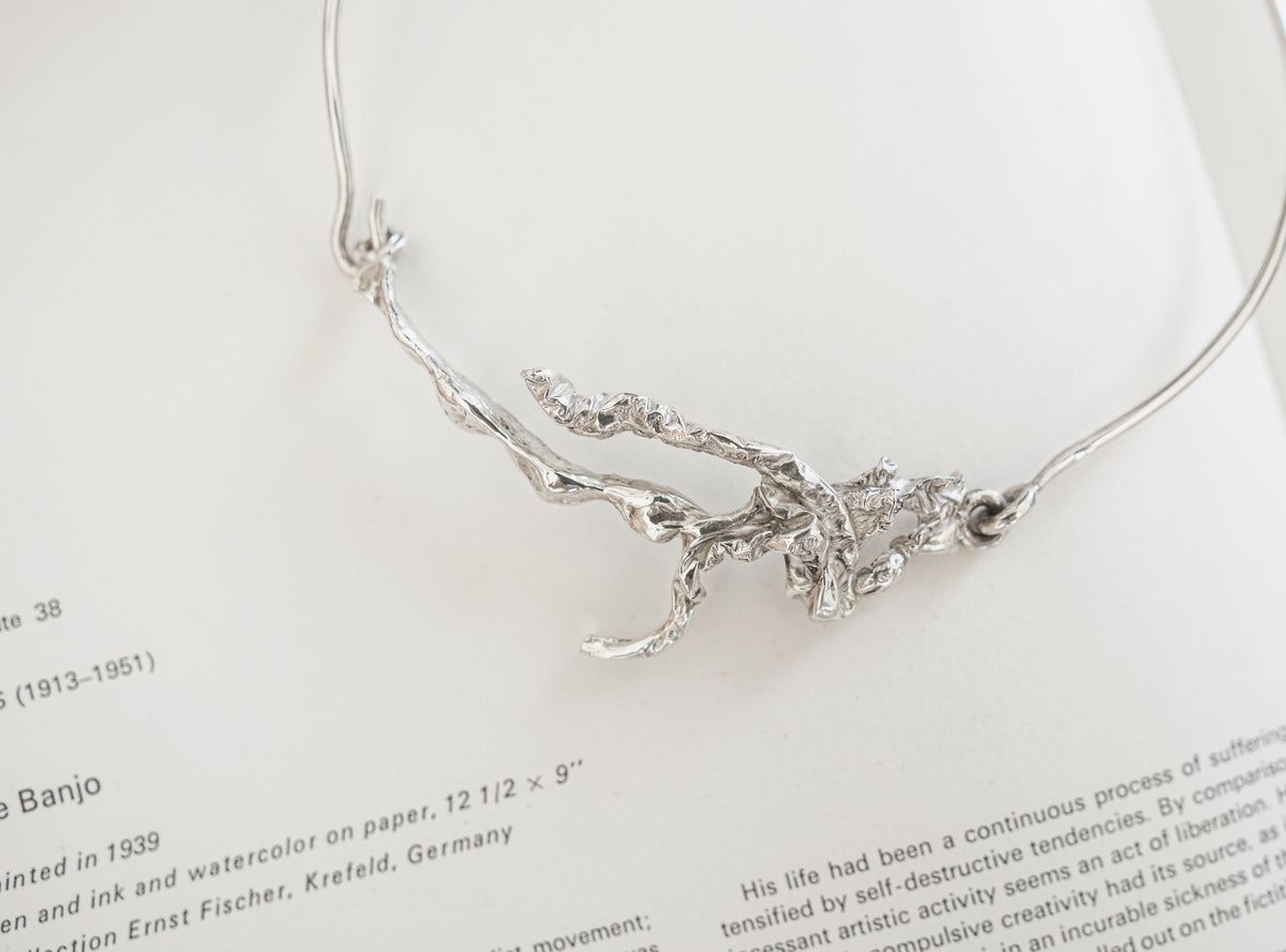 A silver abstract branch or tree-of-life style pendant necklace laid on an open book page with printed text visible.